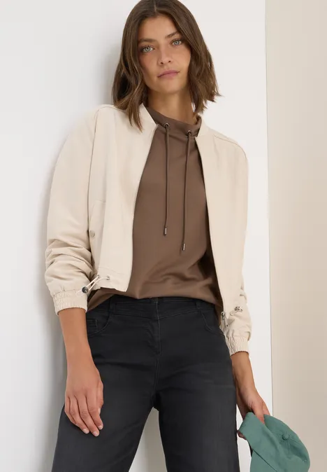 Frau in beiger Jacke, braunem Hoodie und schwarzer Hose steht lässig an einer Wand.